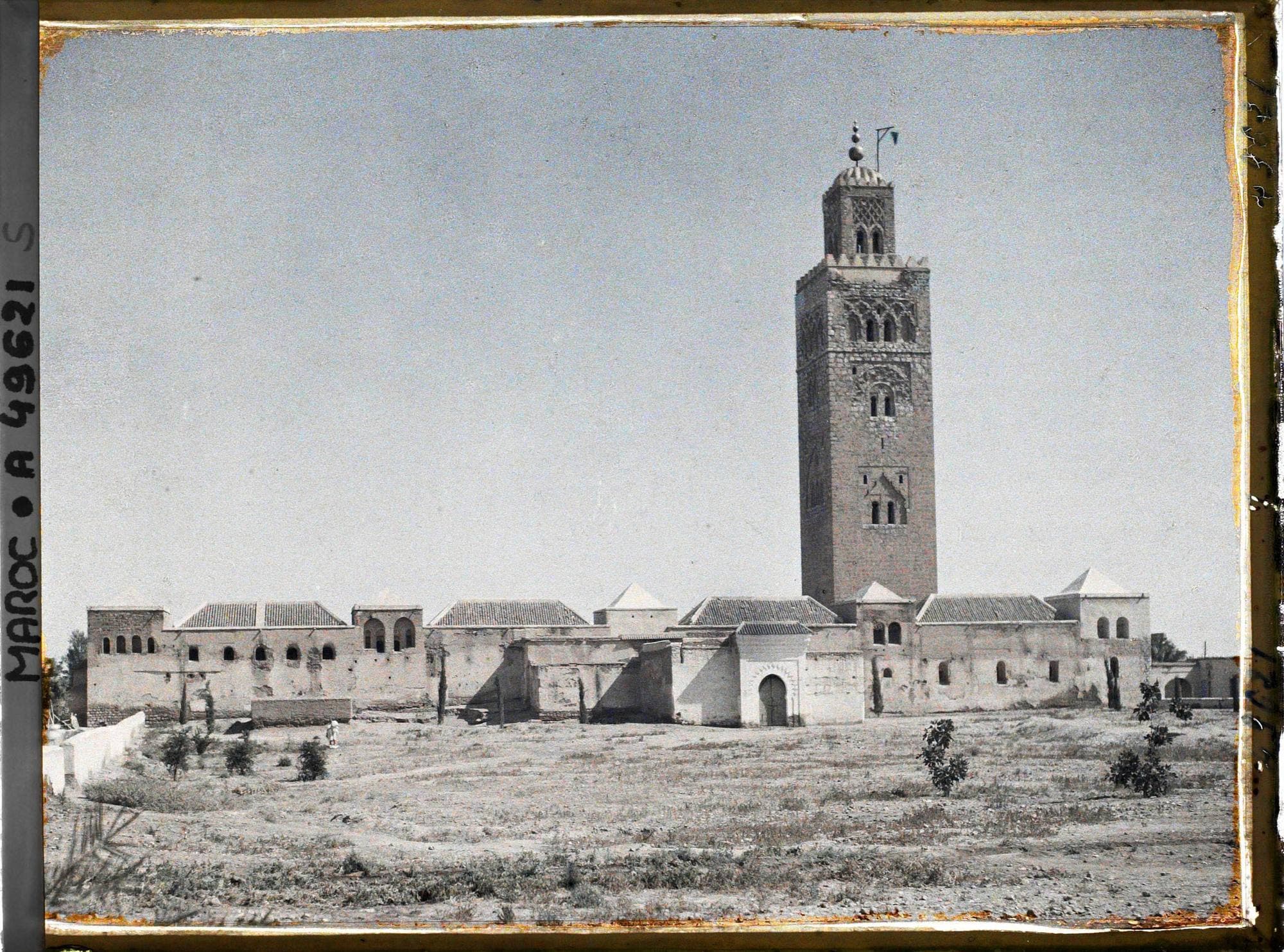 La mosquée Koutoubia vue de face : minaret carré dominant des bâtiments blancs aux toits de tuiles, sur un terrain nu.