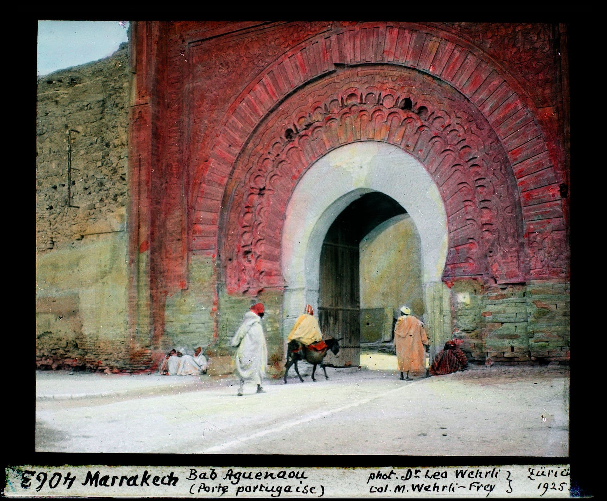 Le grand arc de Bab Agnaou au décor sculpté rouge, avec des passants et un âne devant l'entrée.