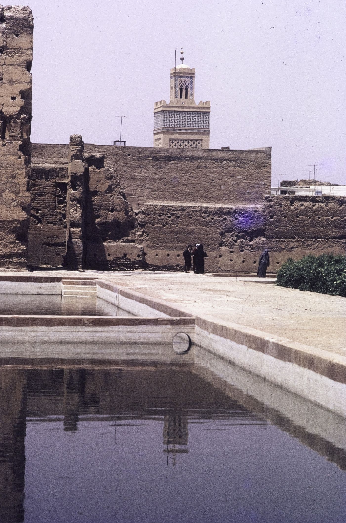Bassin reflétant un mur de pisé, avec le minaret carré d'une mosquée en arrière-plan et quelques silhouettes.