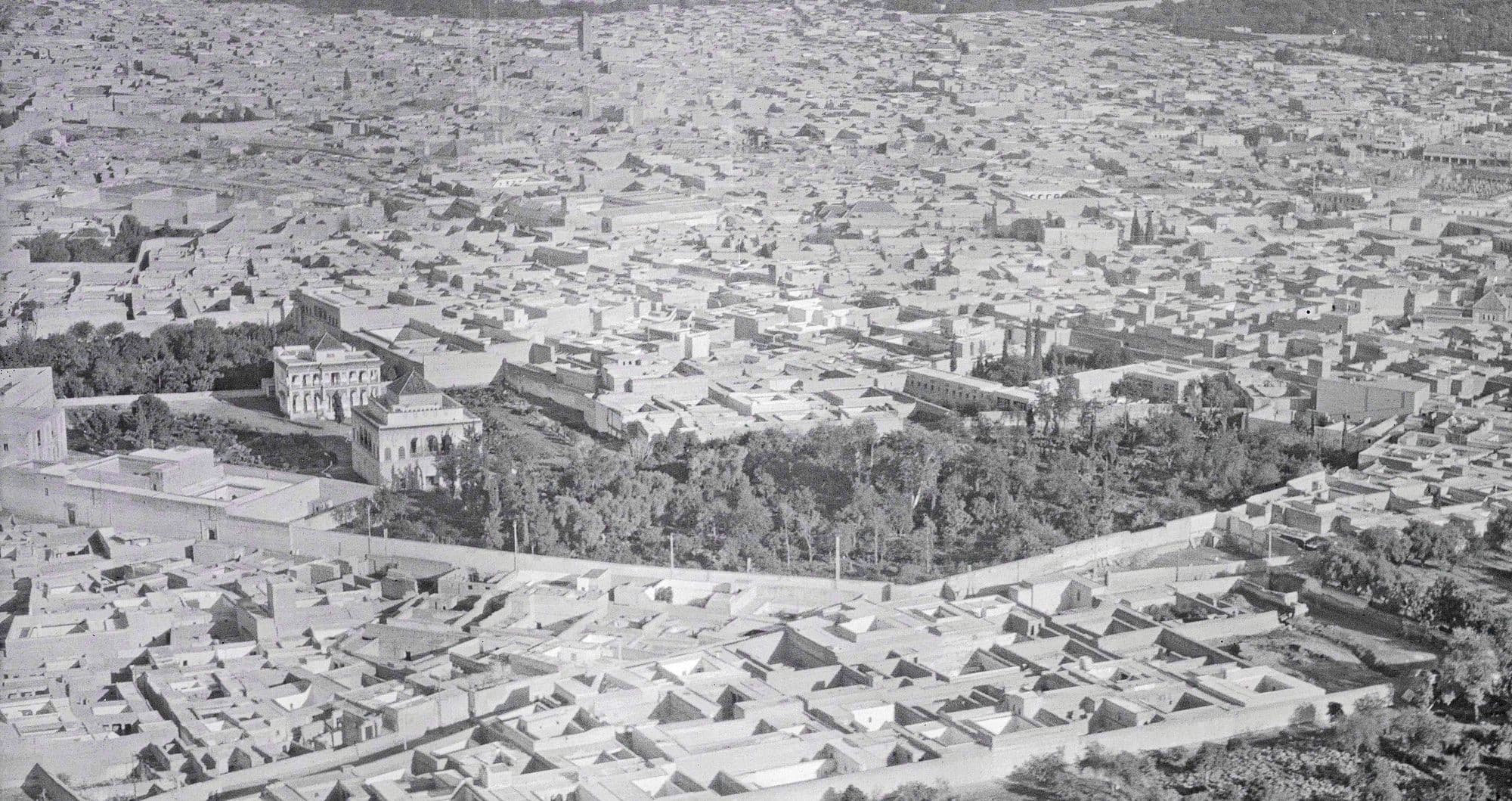 Vue aérienne en noir et blanc de toits-terrasses, avec un bâtiment à toit pyramidal près d'un jardin planté d'arbres.