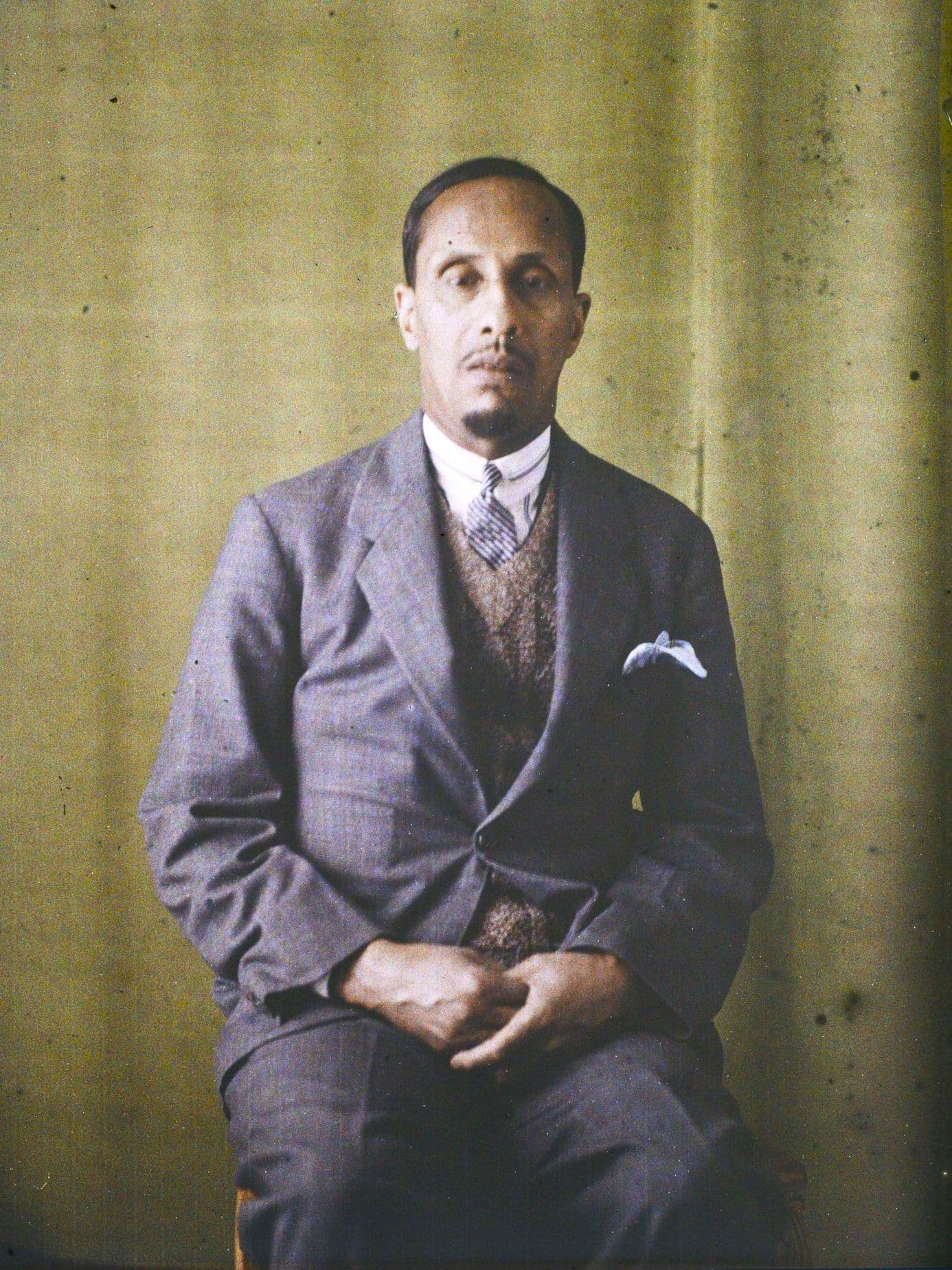 Portrait d'un homme assis en costume, devant un fond uni.