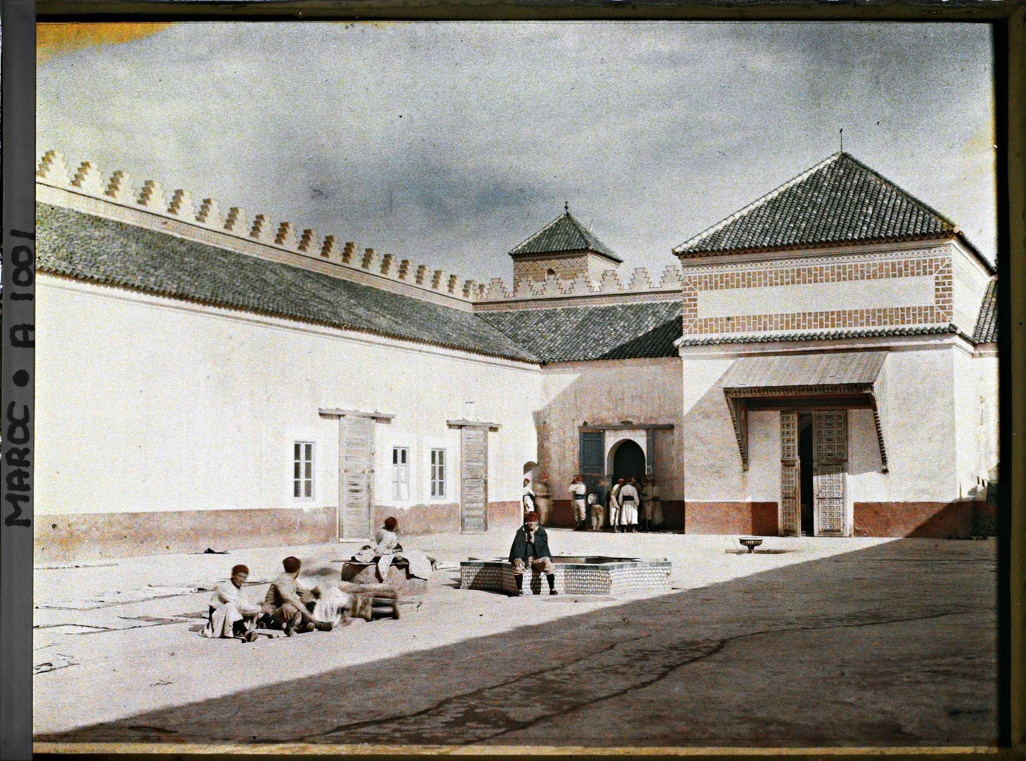 Cour d'un bâtiment à toits de tuiles, avec un bassin au centre et des hommes assis au soleil.