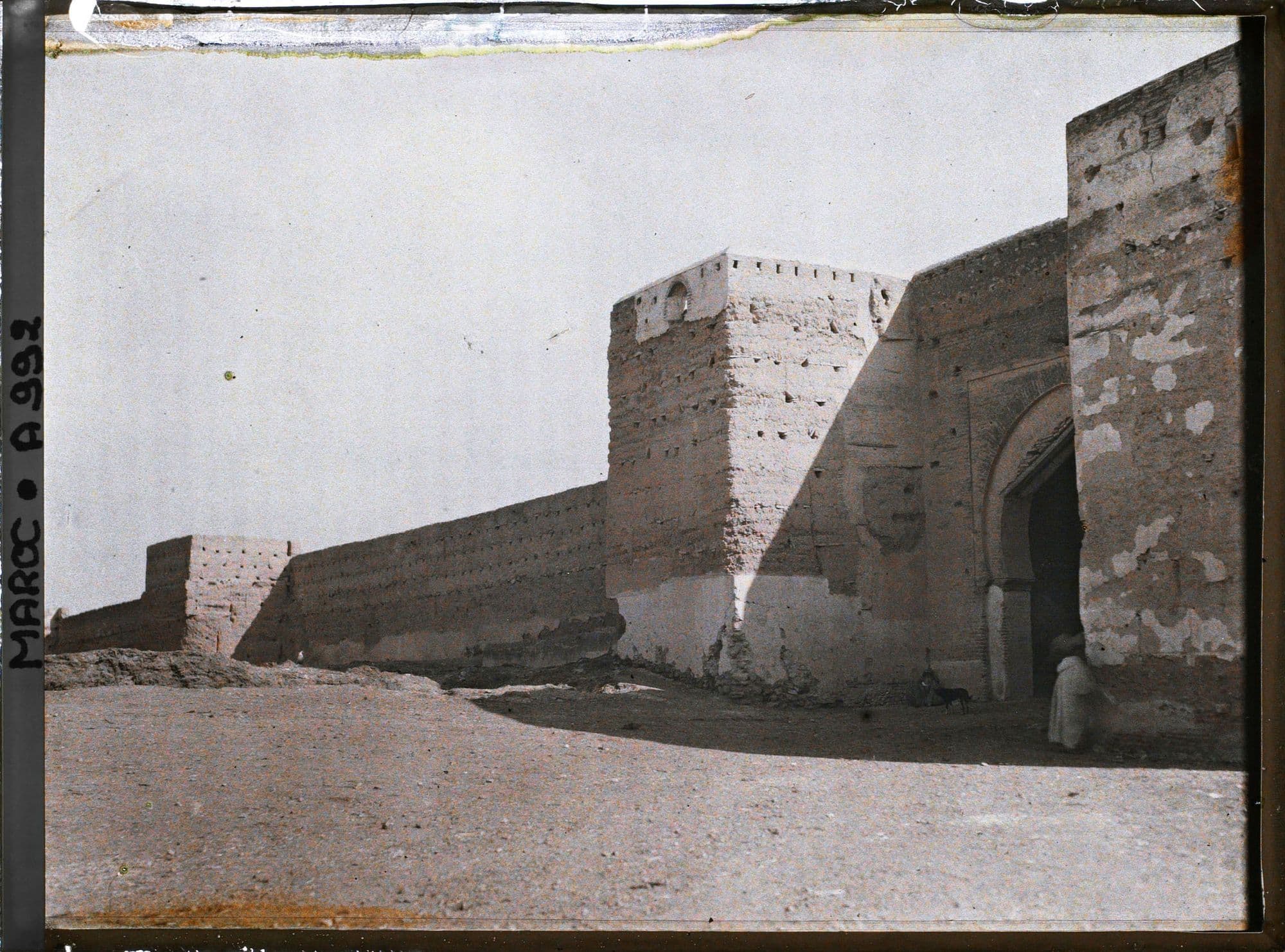 Rempart de pisé flanqué d'une tour carrée et d'une porte en arc, avec un passant devant l'entrée.