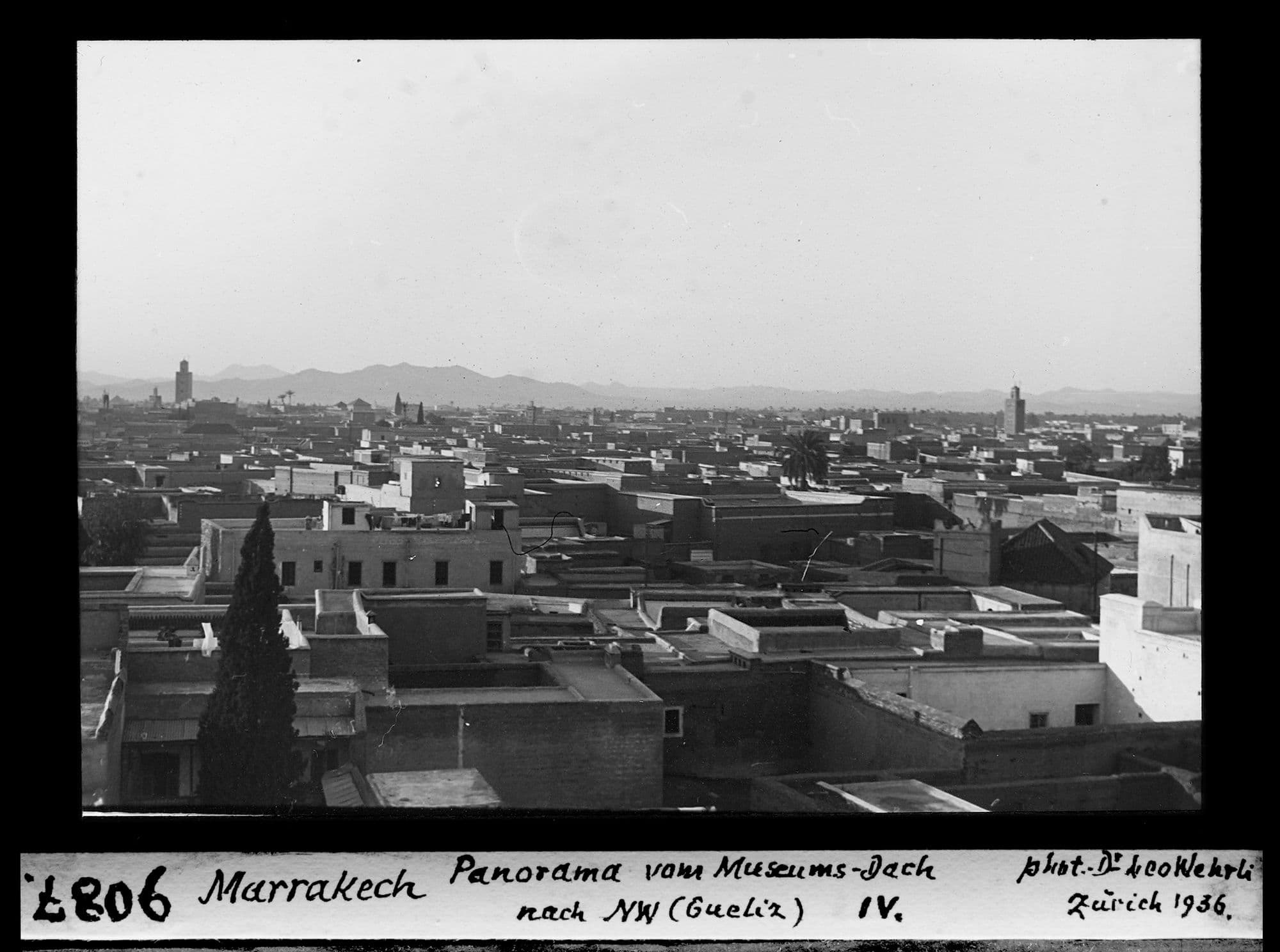 Panorama en noir et blanc sur des toits-terrasses, avec deux minarets et la chaîne de l'Atlas à l'horizon.