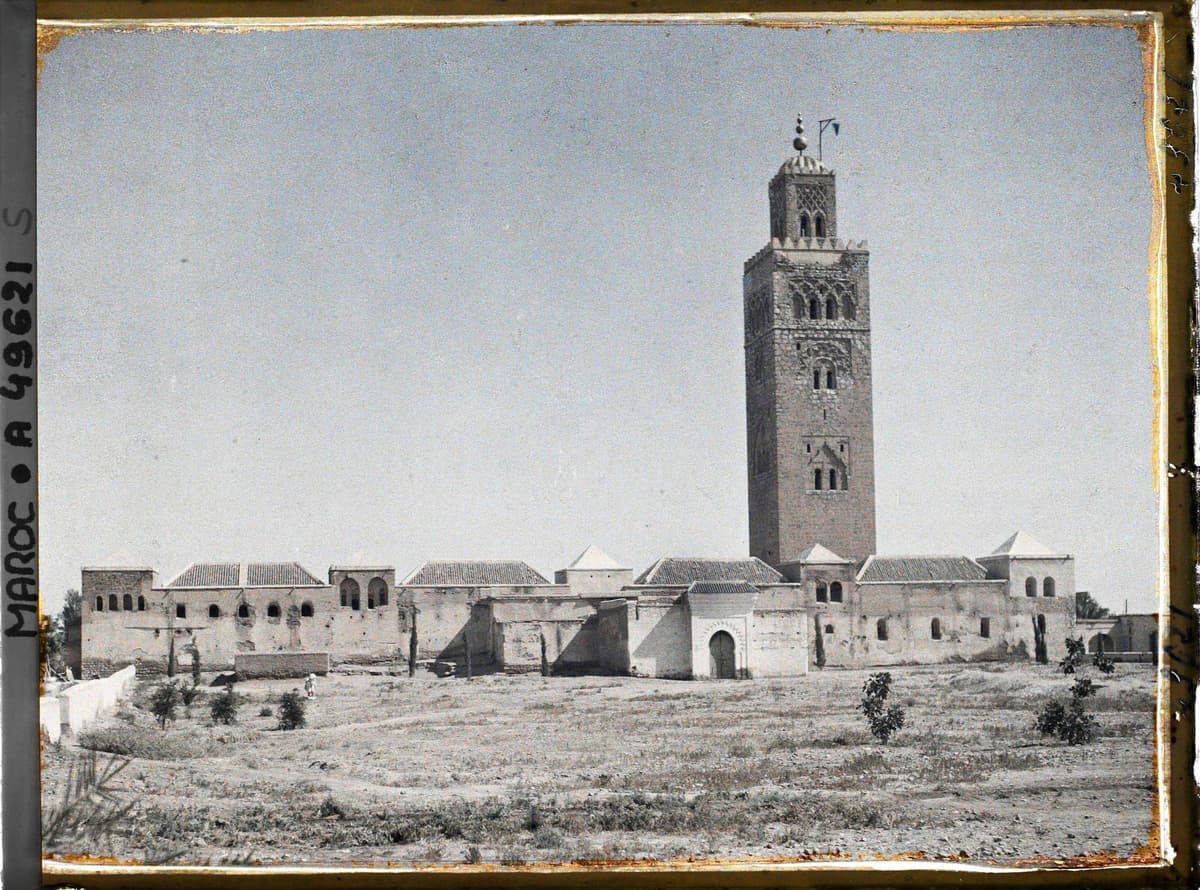 La mosquée Koutoubia vue de face : minaret carré dominant des bâtiments blancs aux toits de tuiles, sur un terrain nu.