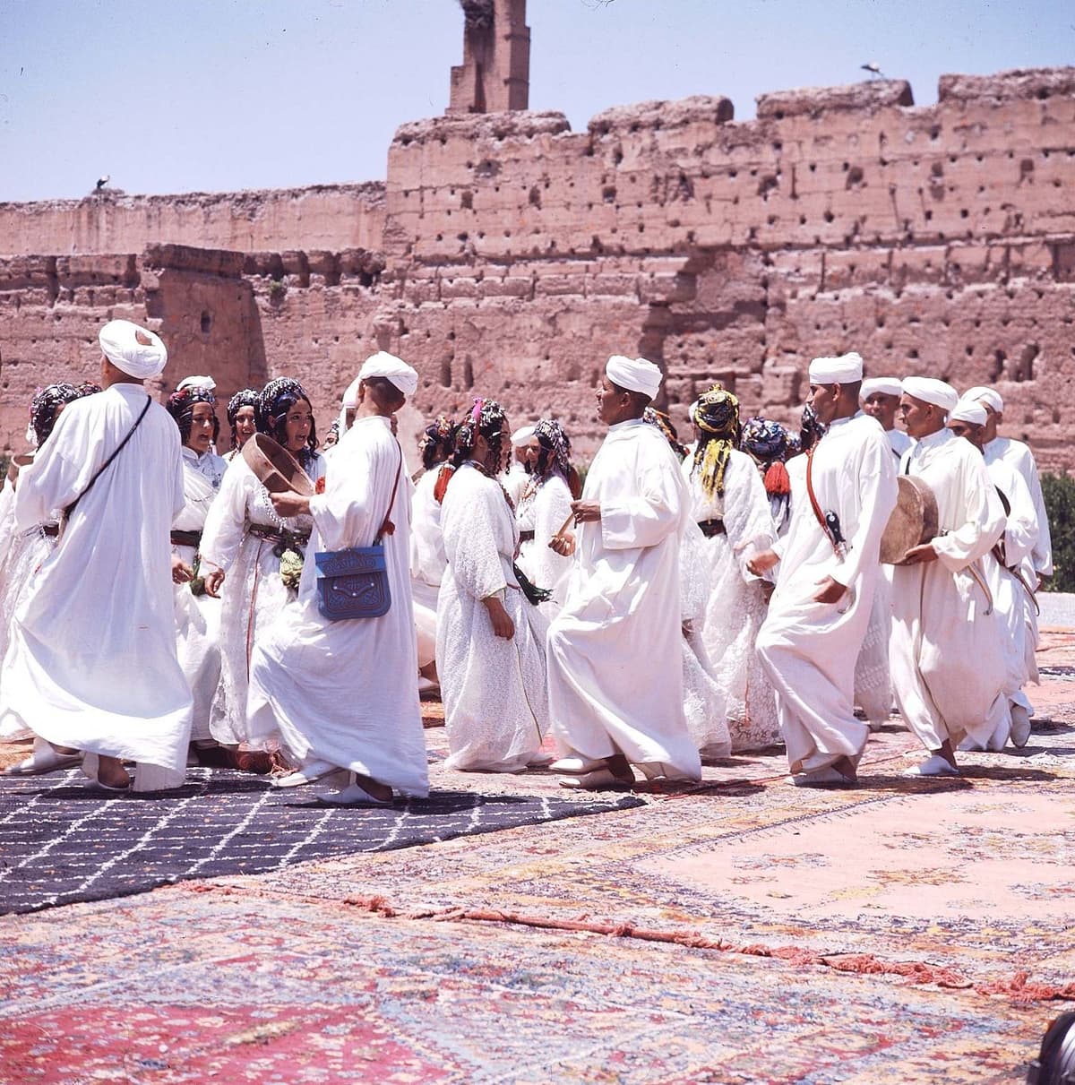 Danseurs et danseuses en tenues blanches sur des tapis étalés, devant de hauts murs de pisé.