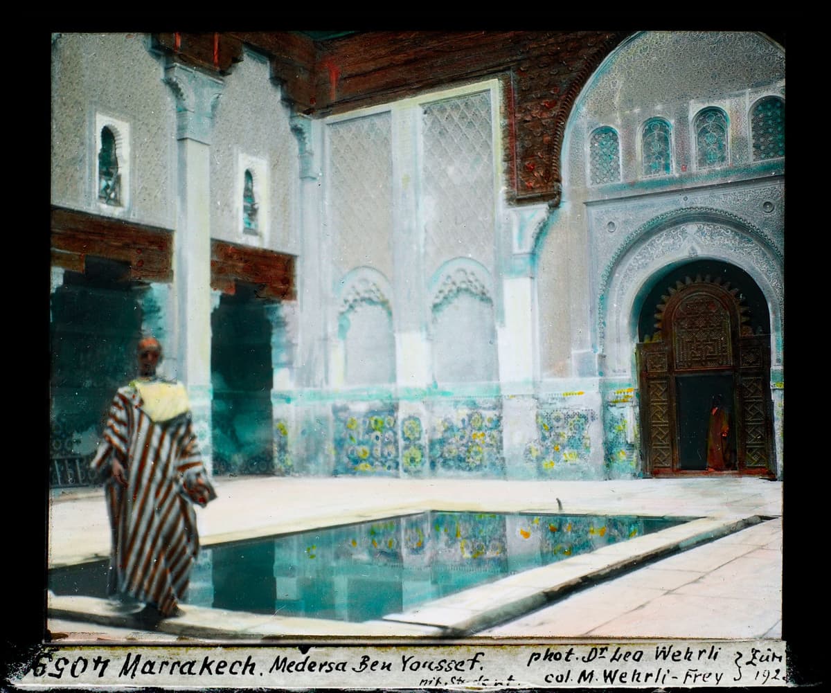 Cour d'une medersa : bassin rectangulaire, façade de stuc ciselé, porte de cèdre sculpté et homme en robe rayée.