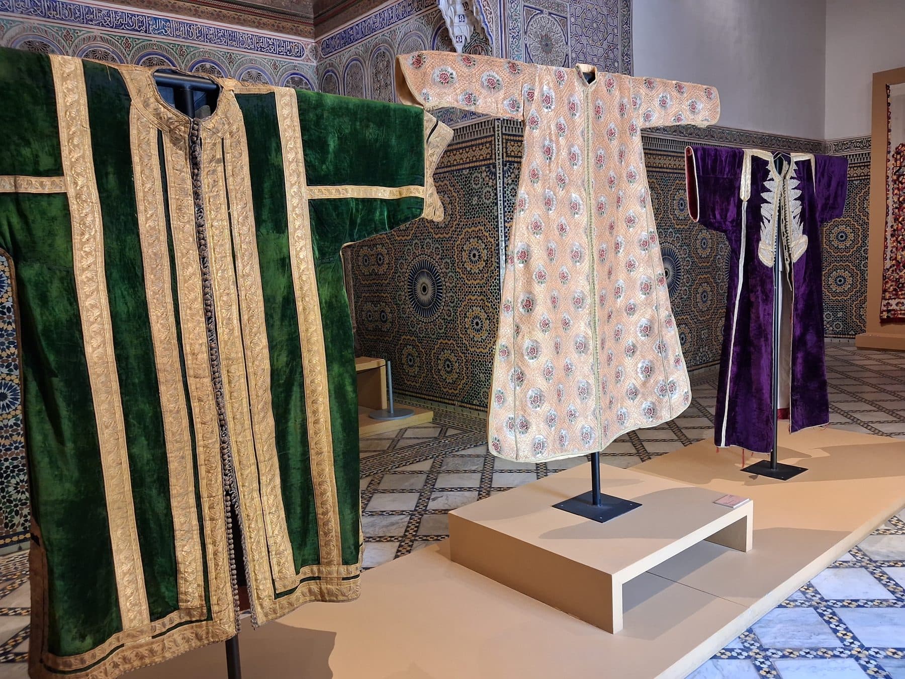 Trois caftans de velours et de broderies exposés devant un mur de zellige, Dar El Bacha