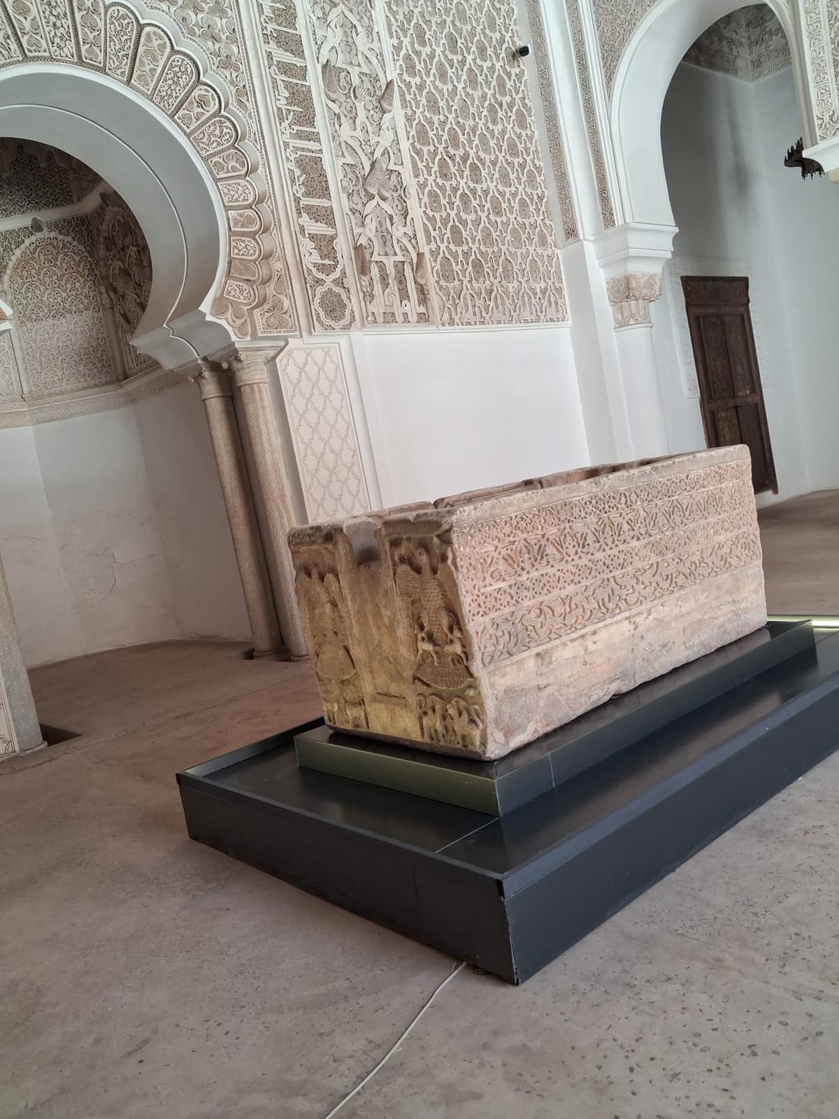 Salle de prière de la medersa Ben Youssef avec son mihrab en arc outrepassé et une vasque de pierre sculptée exposée.