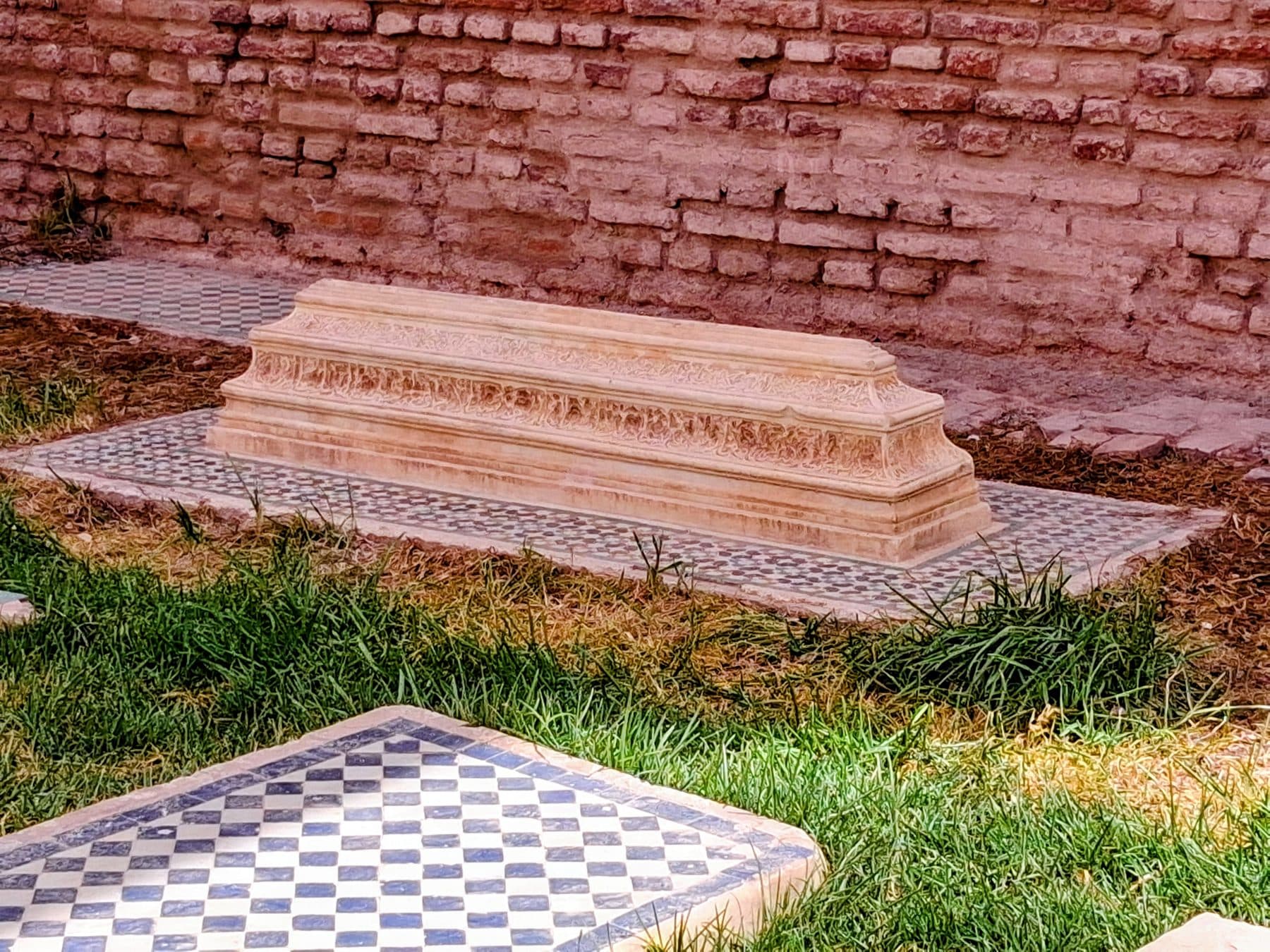 Tombe de marbre sculpté dans l'herbe du jardin, entourée de carreaux de zellige, devant un mur de brique